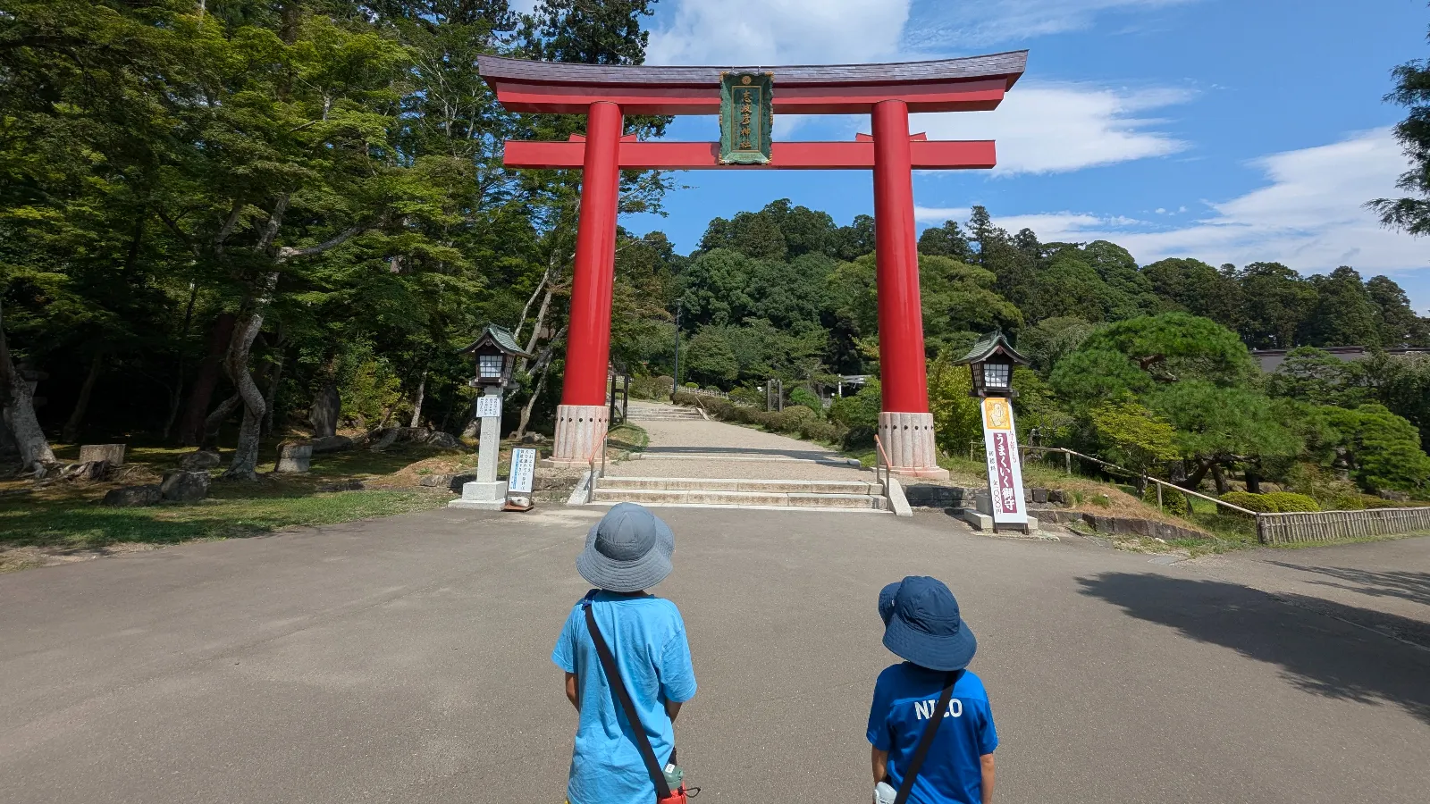 Two children approach the bright red torii gate at Shiogama Shrine in Sendai, Miyagi, Japan under clear summer skies.