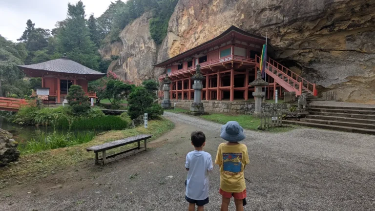 Two children face the red bridge and cliffside Takkoku-no-iwaya Bishamondō hall surrounded by trees and stone lanterns in Hiraizumi, Iwate.