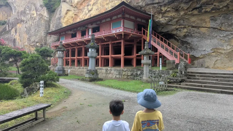 Two children look toward the red cliffside hall of Takkoku-no-iwaya Bishamondō at Takkoku Seiko-ji Temple in Iwate, Japan.
