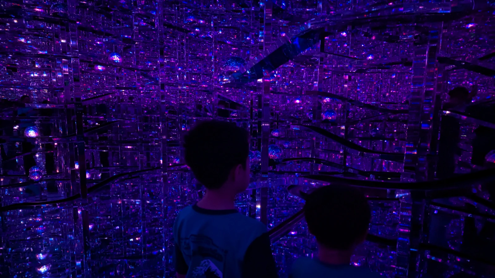Children looking at the glowing glass orbs and mirrored pathways inside the Microcosmos installation at teamLab Borderless Tokyo.