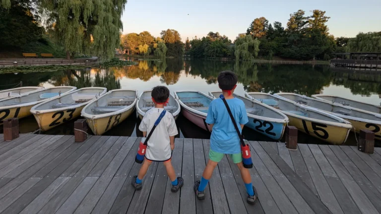 Two boys stand by the wooden dock at Tokiwa Park in Asahikawa, gazing at rows of boats on the calm pond during sunset.