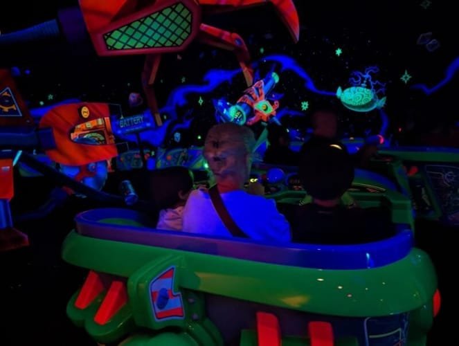 A vibrant scene from Tokyo Disneyland's Buzz Lightyear's Astro Blaster ride. The image shows a green ride vehicle carrying Luca & Nico, and grama illuminated by glowing neon colors of the galactic-themed surroundings, including a large red robotic claw and floating asteroid graphics, creating an exciting intergalactic adventure atmosphere.