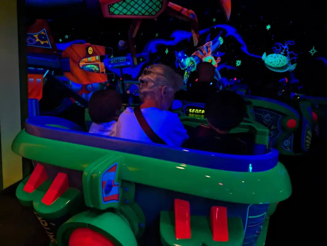 A vibrant scene from Tokyo Disneyland's Buzz Lightyear's Astro Blaster ride. The image shows a green ride vehicle carrying Luca & Nico, and grama illuminated by glowing neon colors of the galactic-themed surroundings, including a large red robotic claw and floating asteroid graphics, creating an exciting intergalactic adventure atmosphere.