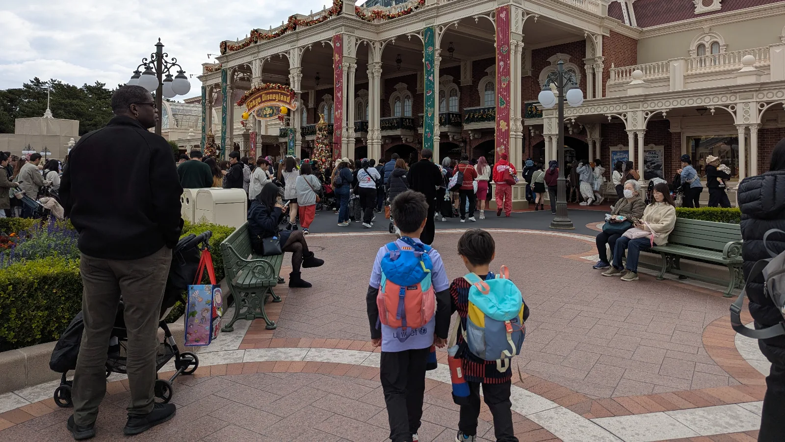 Two kids with backpacks walking toward the Christmas-decorated entrance of Tokyo Disneyland, with crowds gathered under the festive archway.