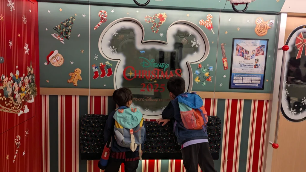 Two kids in backpacks looking at the Mickey-shaped window inside the Tokyo Disney Resort Line Christmas-themed monorail decorated for the 2025 holiday season.