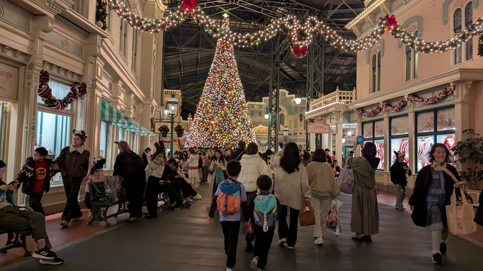 A nighttime view of Tokyo Disneyland’s World Bazaar with a giant Christmas tree glowing above the crowd, while two young kids wearing backpacks walk toward the lights.