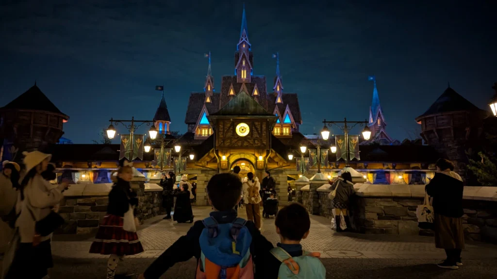 Two children look toward the illuminated Frozen Kingdom castle in Fantasy Springs at Tokyo DisneySea, glowing blue and gold under the night sky.