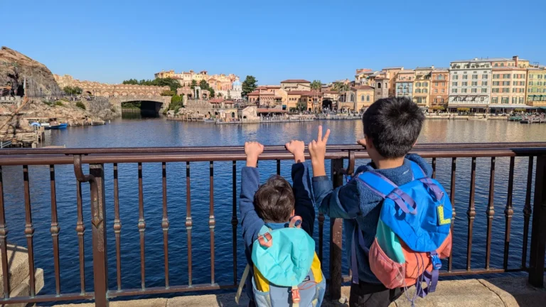 Two children stand at the railing overlooking Tokyo DisneySea’s Mediterranean Harbor, with colorful Italian-style buildings and calm water in front of them.