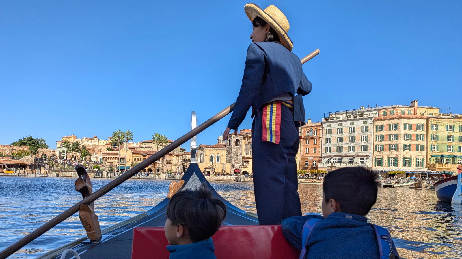 A gondolier rows a Venetian-style gondola at Tokyo DisneySea while two young kids sit at the front, looking toward the Mediterranean Harbor waterfront.