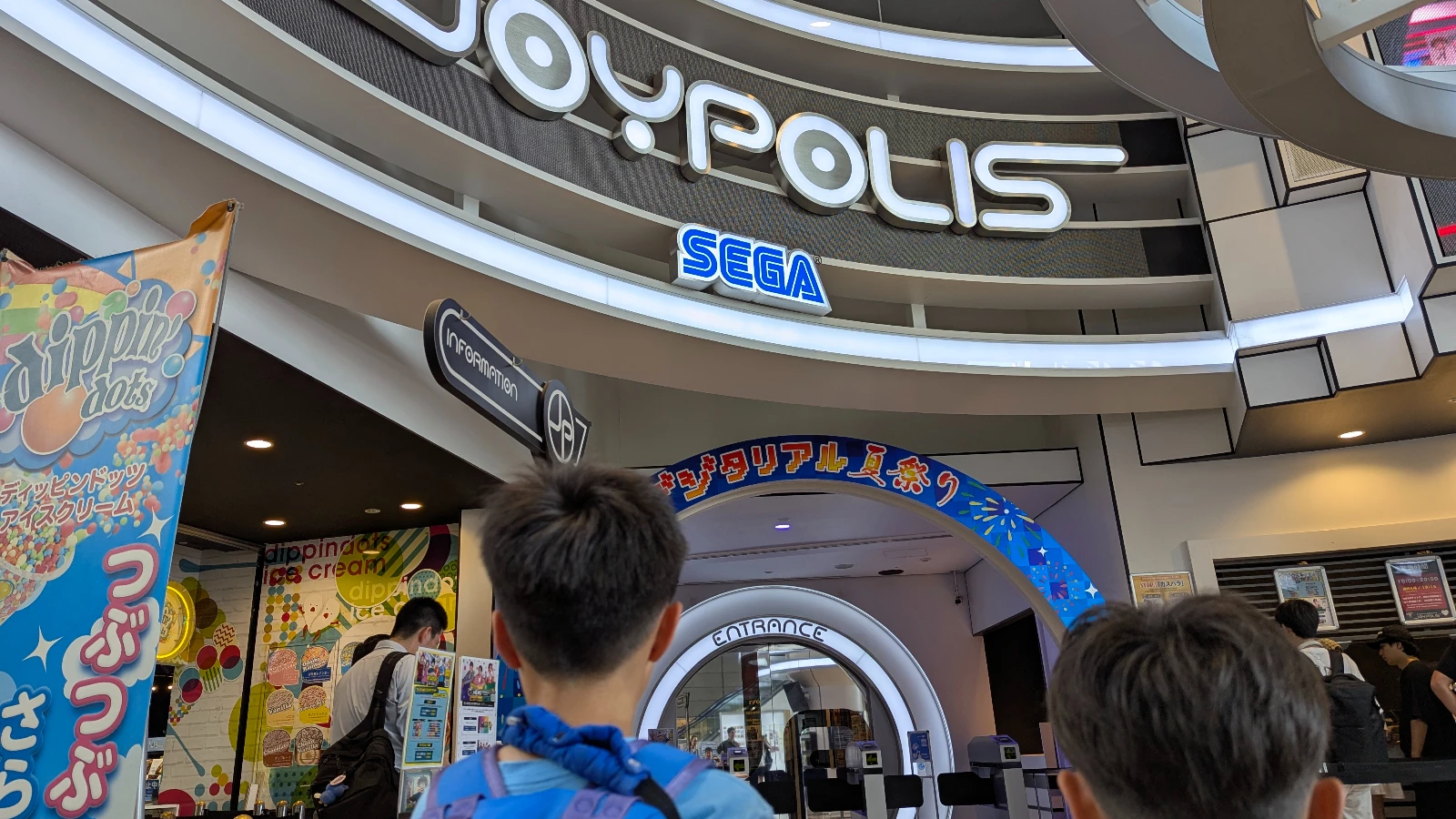 Luca and Nico standing at the entrance of Tokyo Joypolis (東京ジョイポリス) in Odaiba, with the Sega sign above and ticket booths inside.