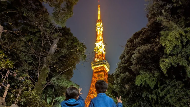 Children looking up at the brightly illuminated Tokyo Tower at night from a park surrounded by trees.