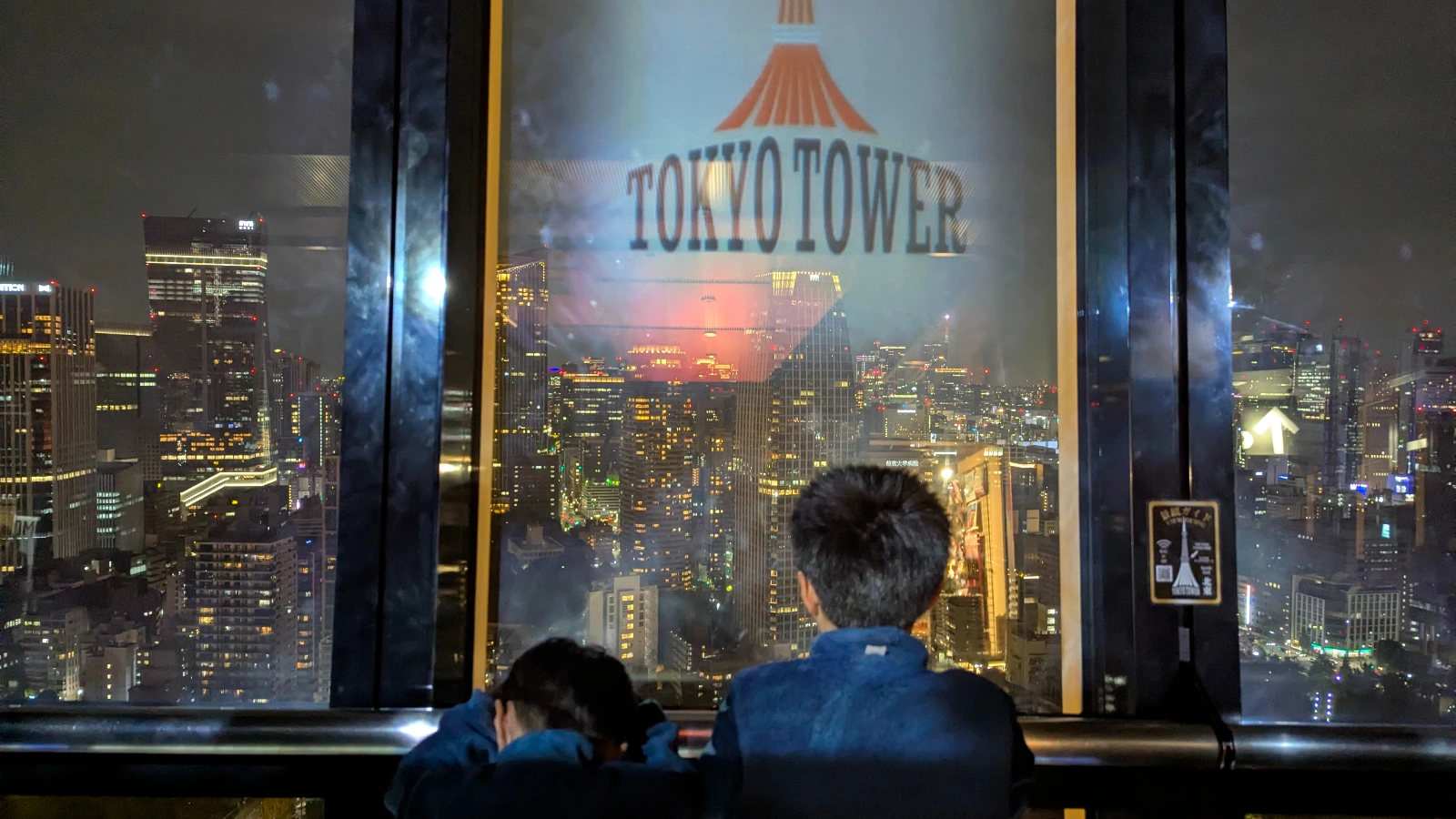 Two children viewing Tokyo’s illuminated skyline from the Tokyo Tower observation deck with the tower logo reflected on the window.