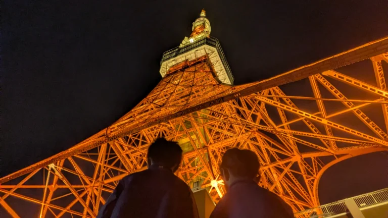 Two children standing beneath Tokyo Tower at night, looking up at the glowing lattice structure from below.
