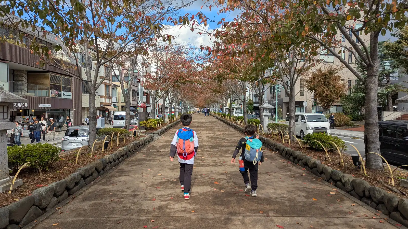 Children walking along the Dankazura pedestrian approach to Tsurugaoka Hachimangu in Kamakura, lined with trees and shops on both sides.