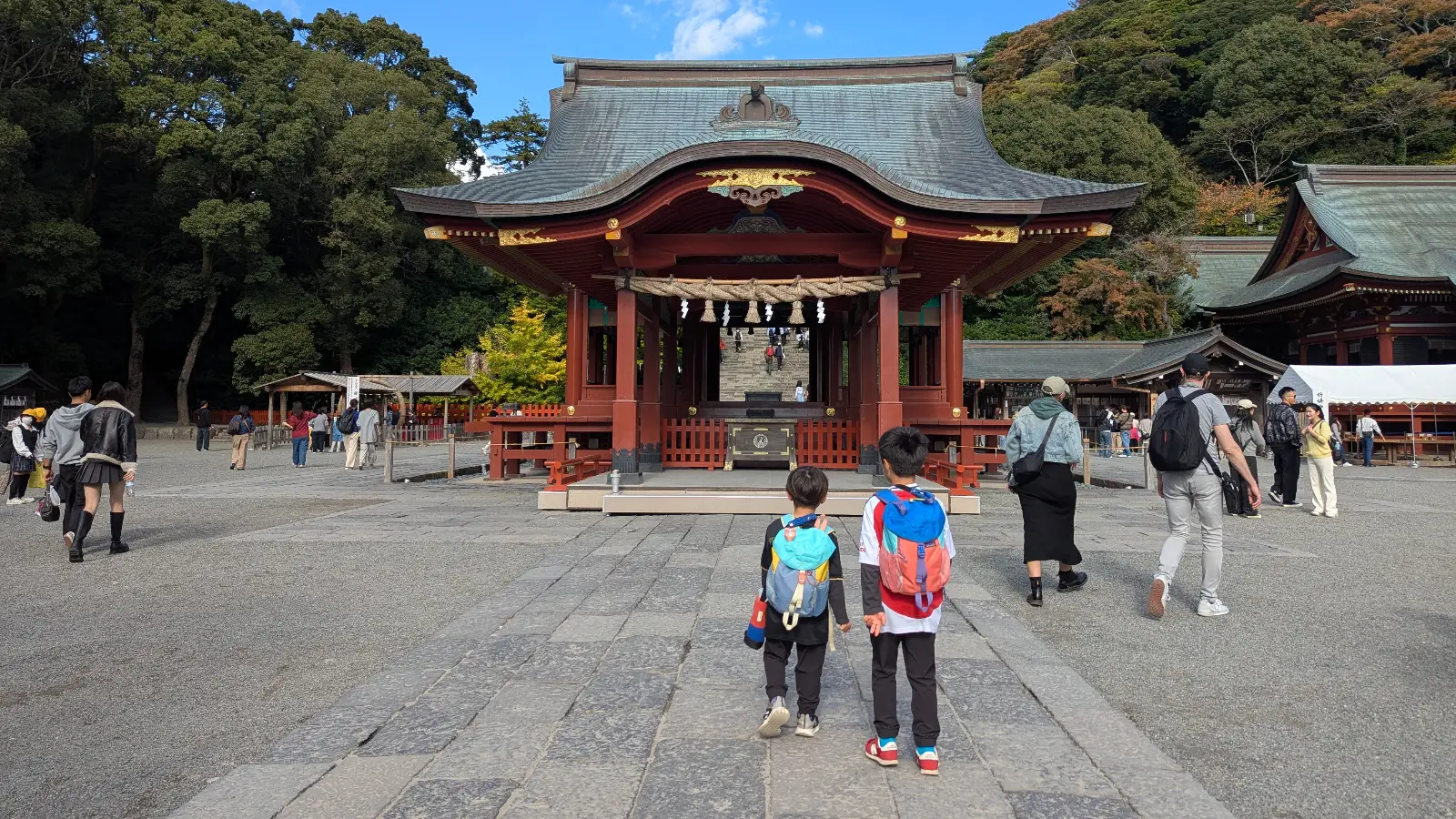 Children walking toward the main hall of Tsurugaoka Hachimangu Shrine in Kamakura, with visitors exploring the courtyard and forested backdrop.