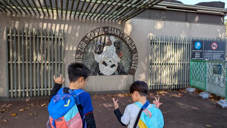 Two kids standing in front of the Tokyo Ueno Zoo Giant Panda House mosaic sign, making playful hand gestures during their visit.