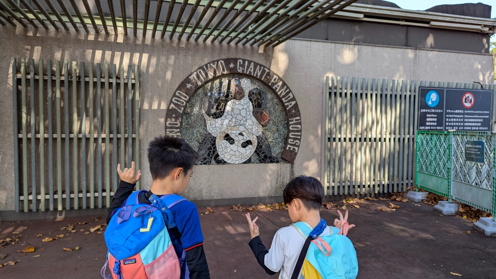 Two kids standing in front of the Tokyo Ueno Zoo Giant Panda House mosaic sign, making playful hand gestures during their visit.