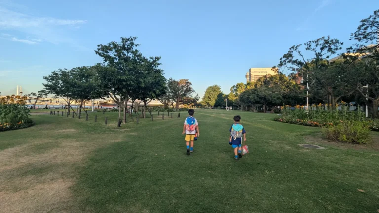 Luca & Nico walking through Yamashita Park in Yokohama on a sunny day, surrounded by green lawns, trees, and flowers with views of the bay in the distance.