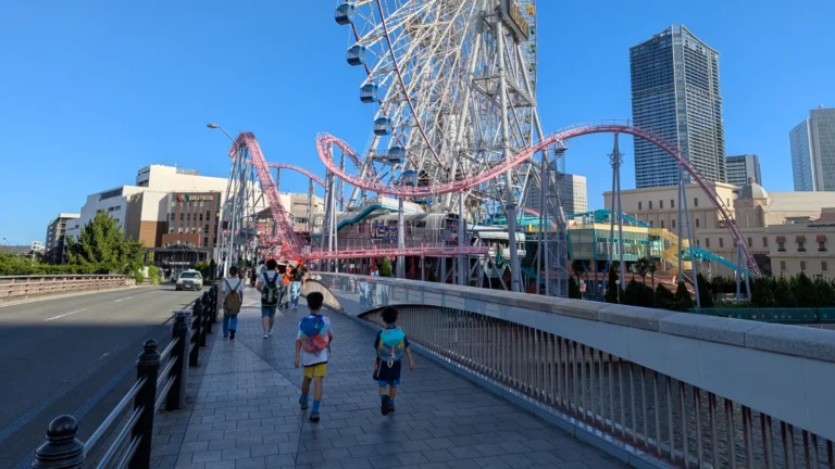 Luca & Nico walking toward Yokohama Cosmo World with the Cosmo Clock 21 Ferris wheel and roller coaster in view under a clear blue sky