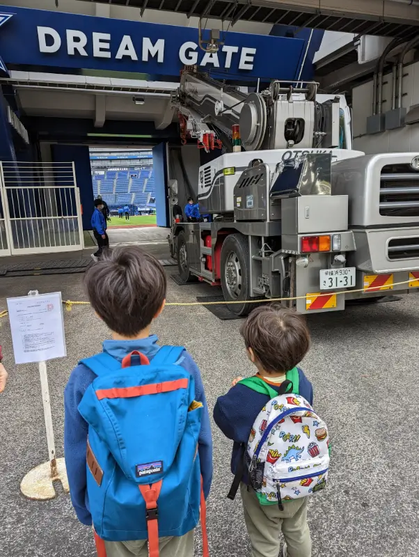 Luca & Nico with backpacks stand in front of Yokohama’s “Dream Gate” stadium entrance, capturing the city’s love for family-friendly sports venues.