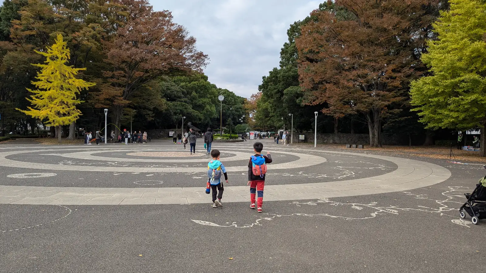Two children walking across the central plaza at Yoyogi Park in Tokyo, surrounded by autumn-colored trees and wide open space.