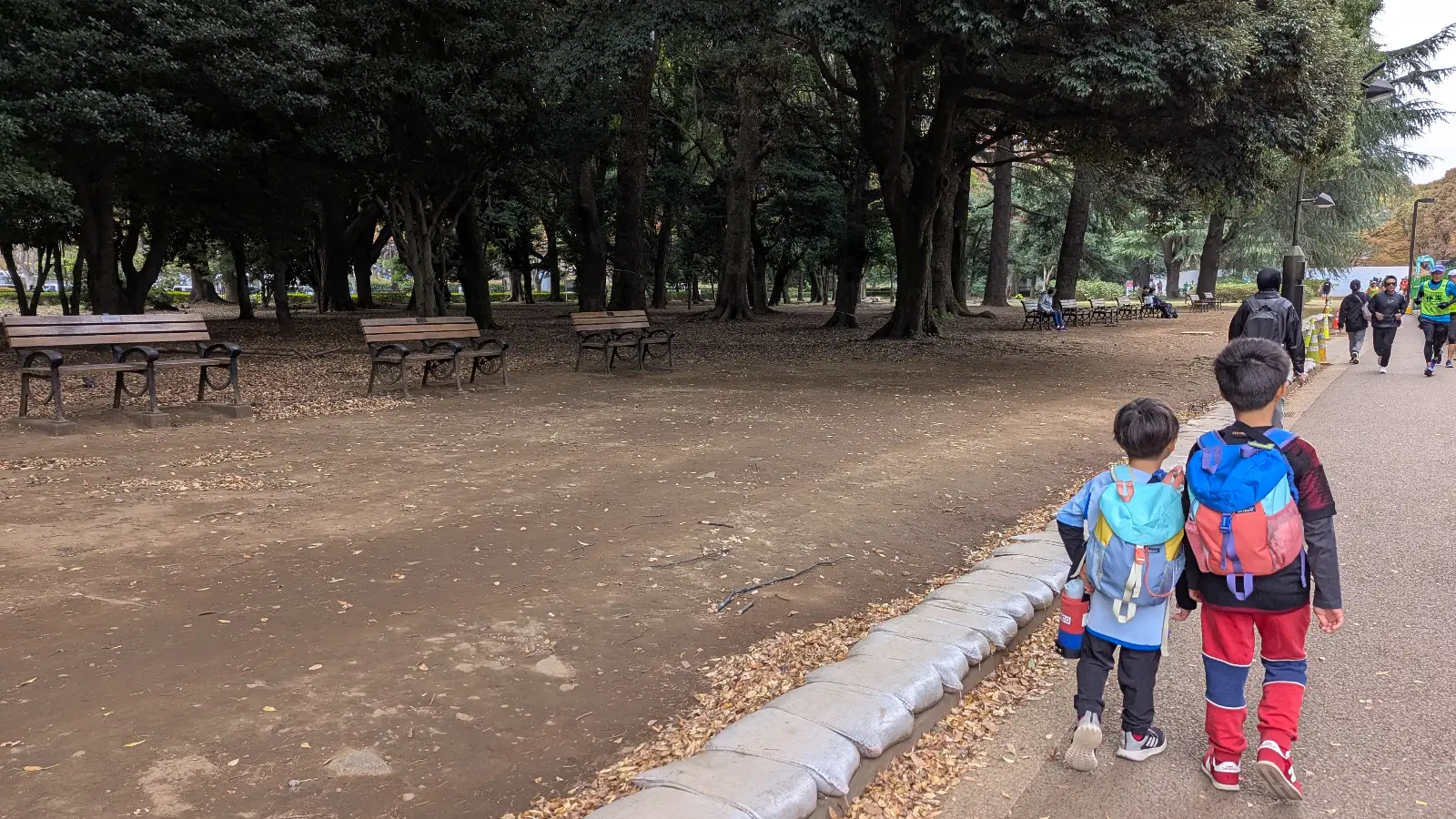Two children walking along a tree-lined path in Yoyogi Park in Tokyo, passing wooden benches and quiet forest areas.